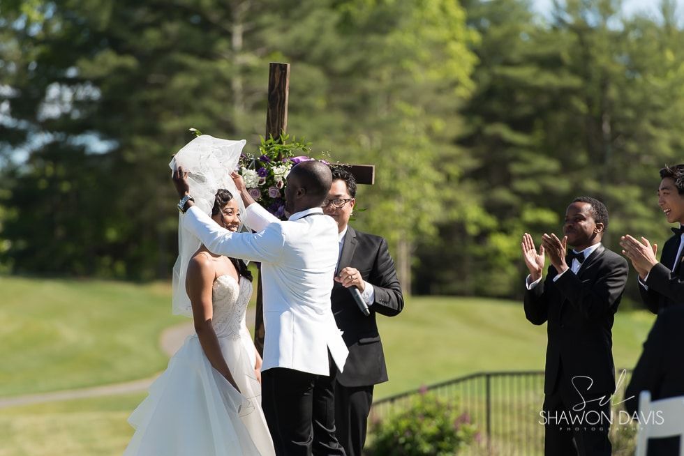 Wedding couple kissing under a canopy outdoors, surrounded by applauding guests.