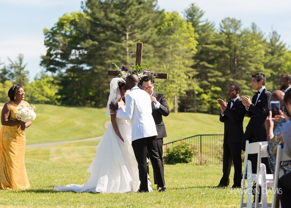 Wedding ceremony outdoors on a grassy lawn, with the bride and groom at center and guests watching nearby