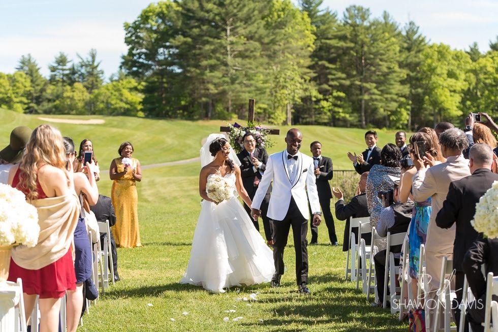 Bride and groom walk down an outdoor aisle as guests applaud at a sunny garden wedding