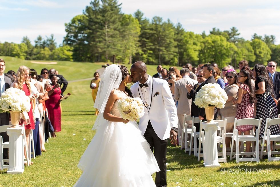 Bride and groom stand at outdoor wedding ceremony in a sunny grassy field, surrounded by seated guests