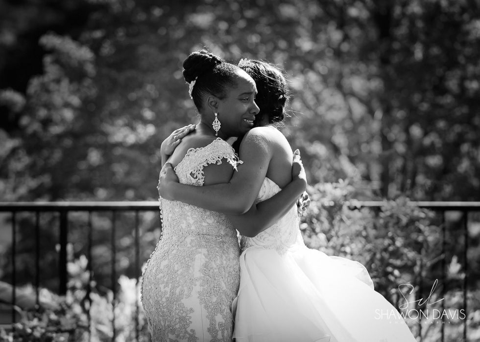 Two people in wedding dresses embracing outdoors in front of a blurred garden background