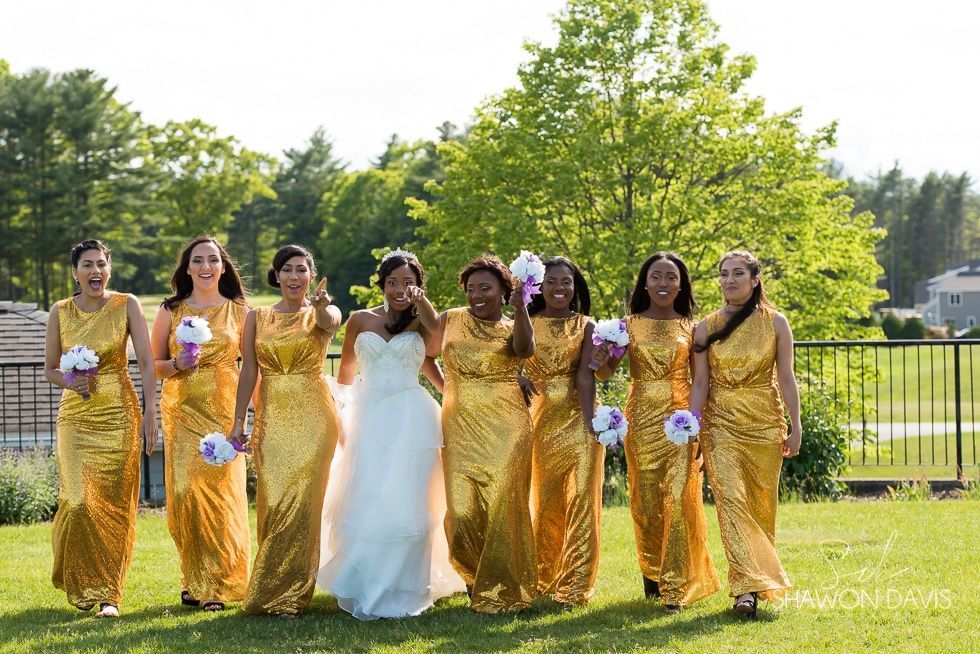 Six women in gold and white dresses pose outdoors on a sunny lawn.