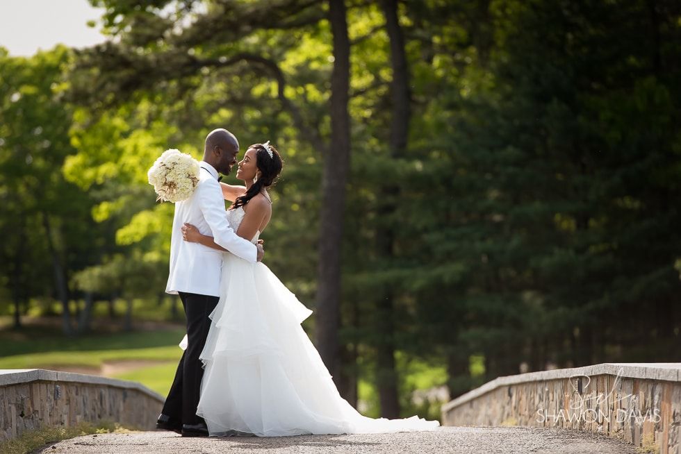 Bride and groom embrace outdoors on a stone bridge in a green park, holding a bouquet