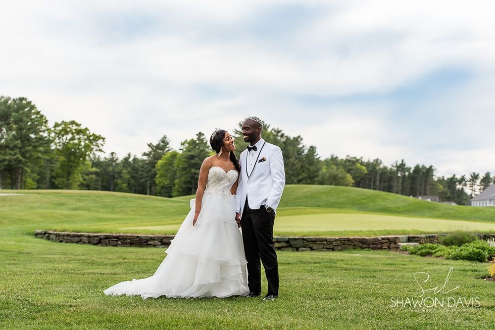 Bride and groom stand in a white gown and tuxedo on a green lawn under a cloudy sky.