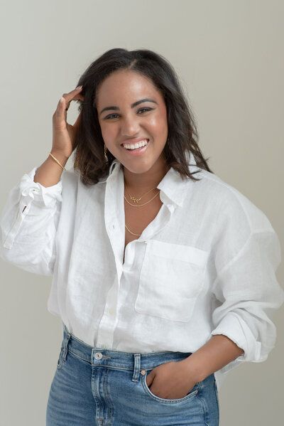 Smiling woman in a white shirt and jeans, standing with hand in pocket against a plain background
