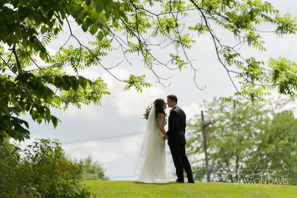 Bride and groom in white and black stand beneath leafy branches in a grassy outdoor setting