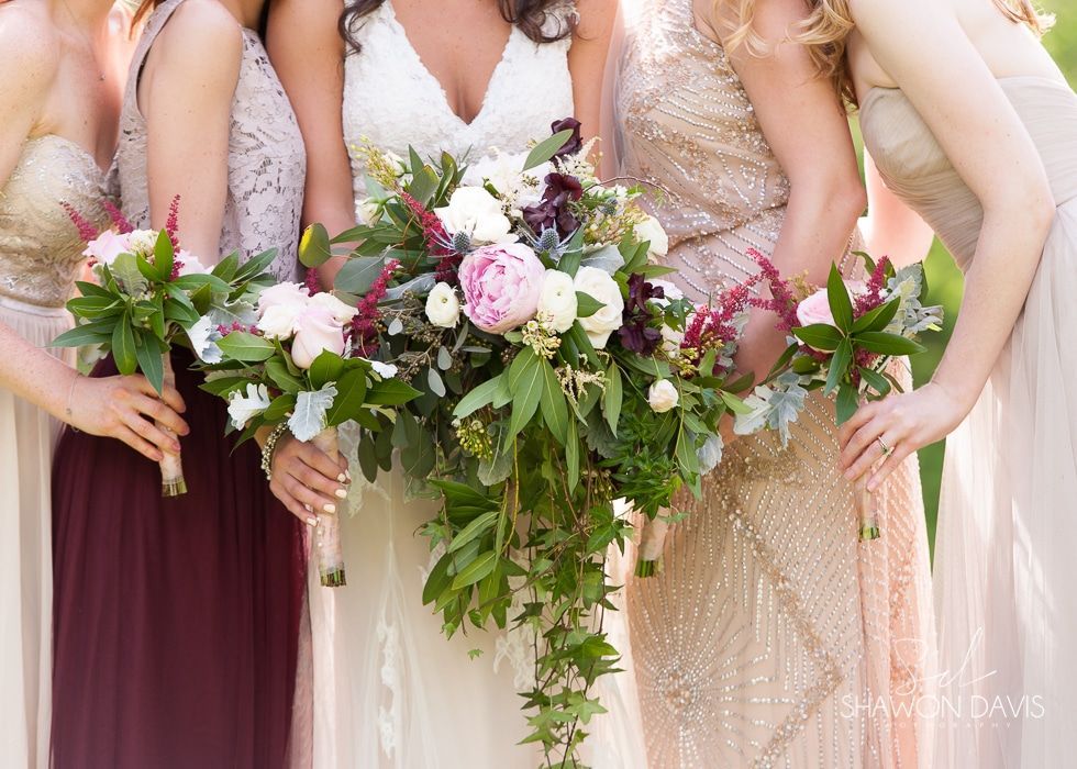 Bridal party holding cascading bouquets in burgundy, blush, white, and gold dresses