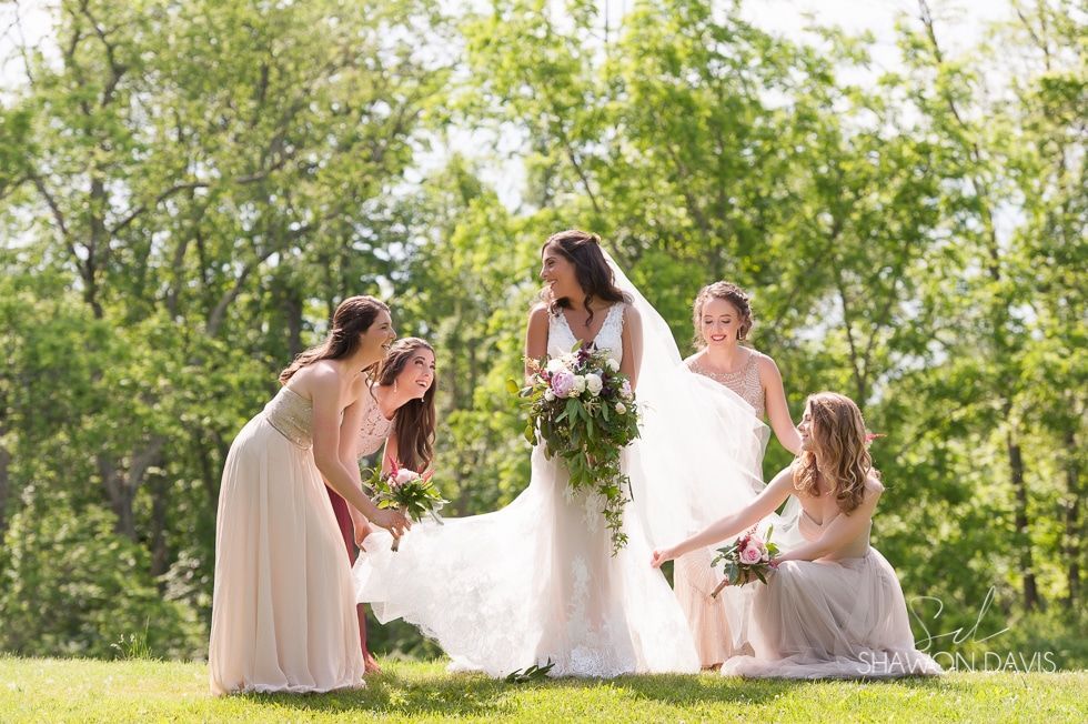 Bridesmaids surround a bride outdoors on a sunny lawn, adjusting her white wedding dress and bouquet.