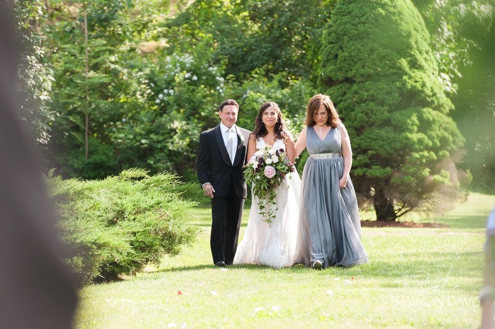 Three people in formal attire walk together on a grassy path, with trees and flowers in a park background.