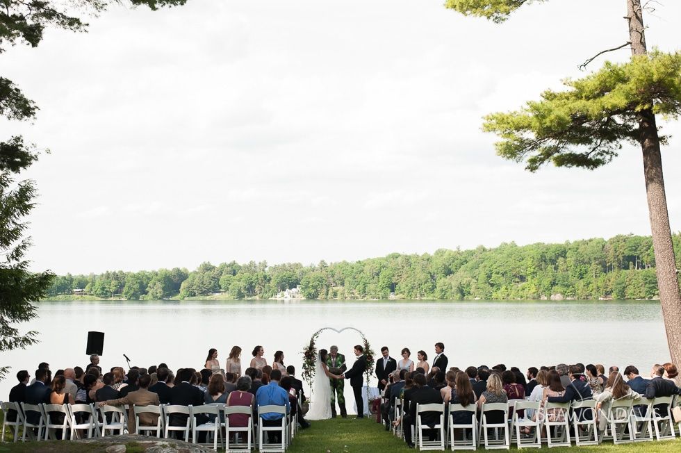 Outdoor wedding ceremony by a lake with guests seated on white chairs under trees