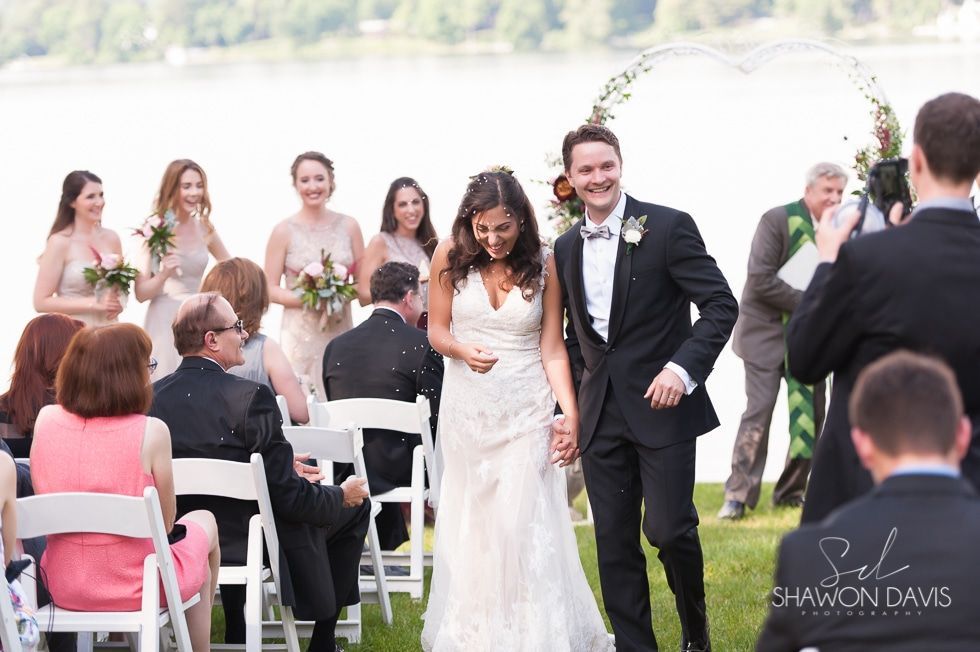 Bride and groom walking down the aisle outdoors, surrounded by guests and bridesmaids holding bouquets.