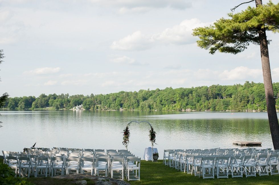 Wedding ceremony setup with white chairs by a calm lakeside under a tree