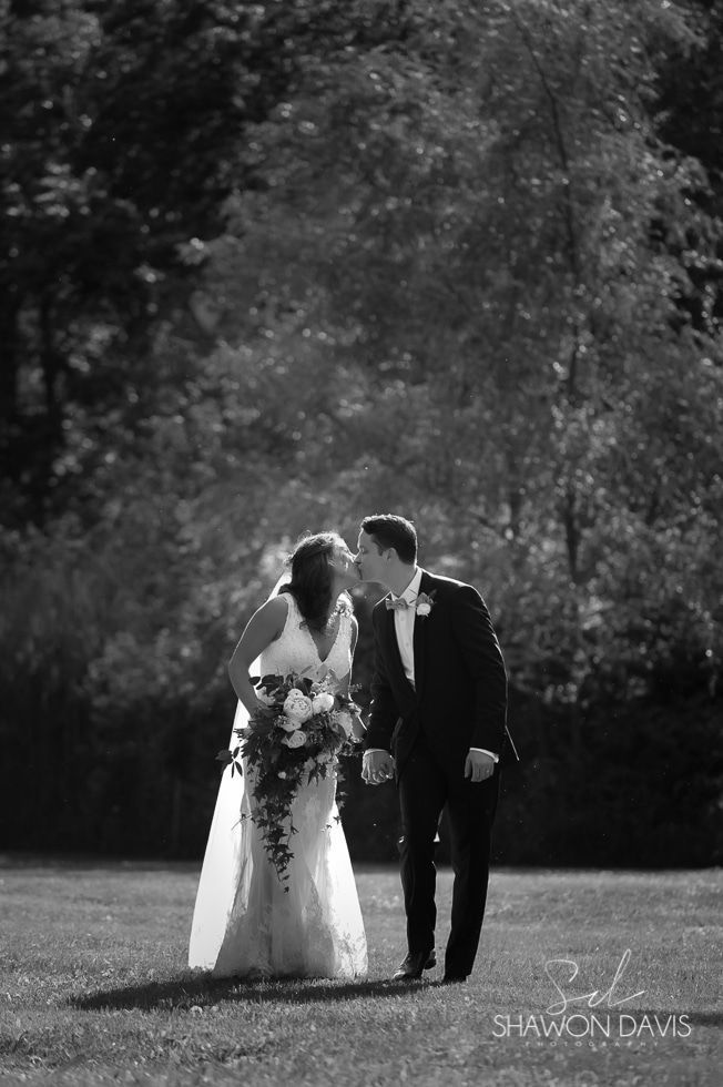 Bride and groom kissing outdoors in black-and-white, with trees and bright sunlight in the background