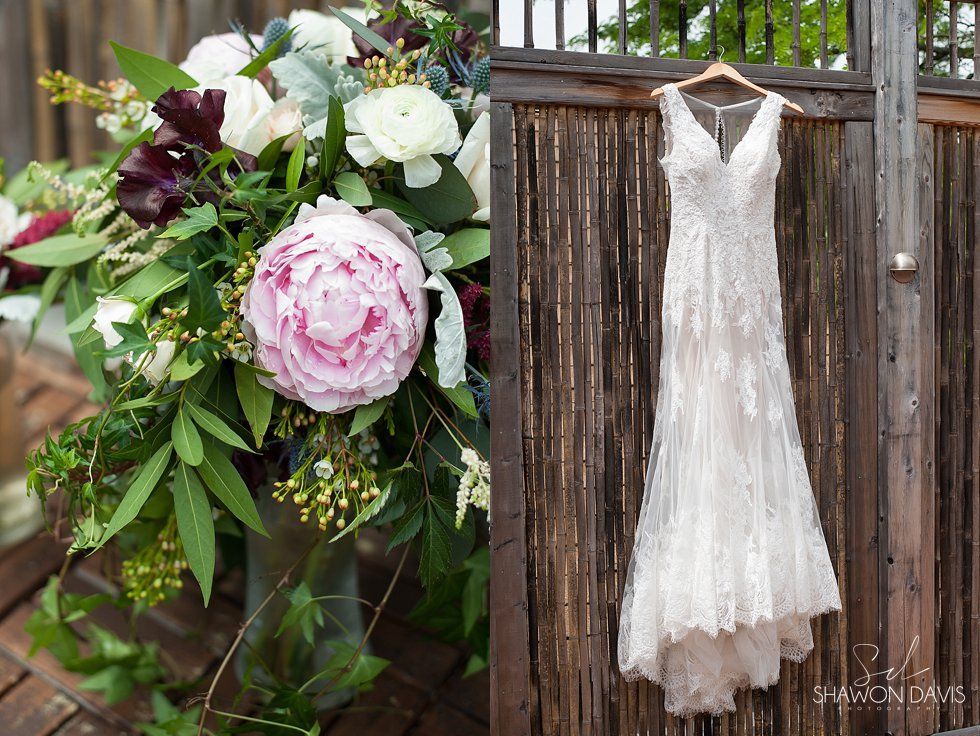 White lace wedding dress hanging beside a pink-and-white floral bouquet on a wooden fence outdoors
