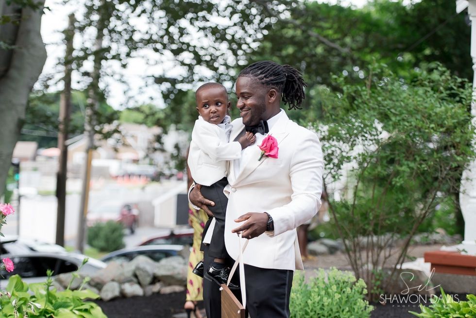 Person holding a child outdoors in a leafy setting, both smiling and dressed formally