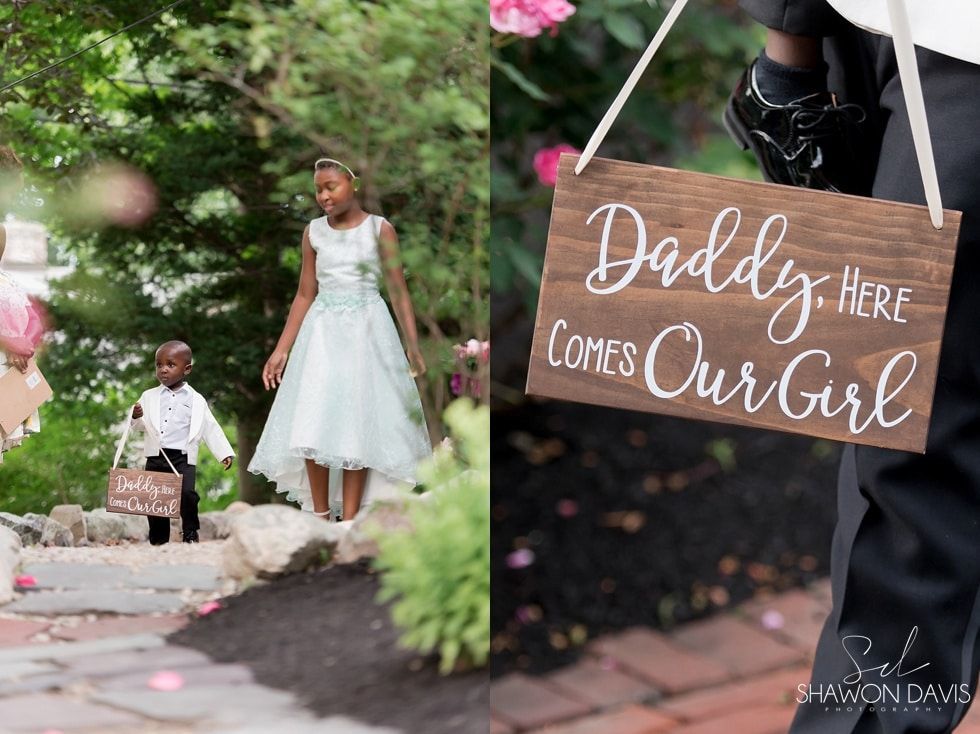 Children in formal attire beside a sign reading “Daddy, Here Comes Our Girl” at an outdoor celebration