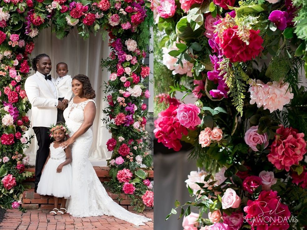Wedding couple and child posing on a flower-covered stage with pink and white floral backdrop.