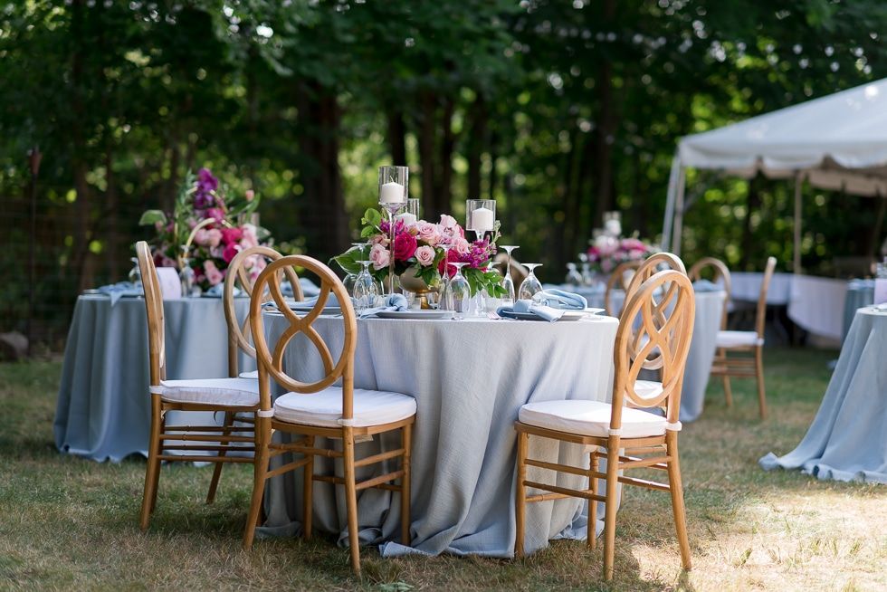 Outdoor wedding reception table with white linens, floral centerpieces, and wooden chairs under trees