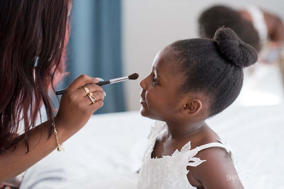 Person applies makeup to a child in a white dress indoors