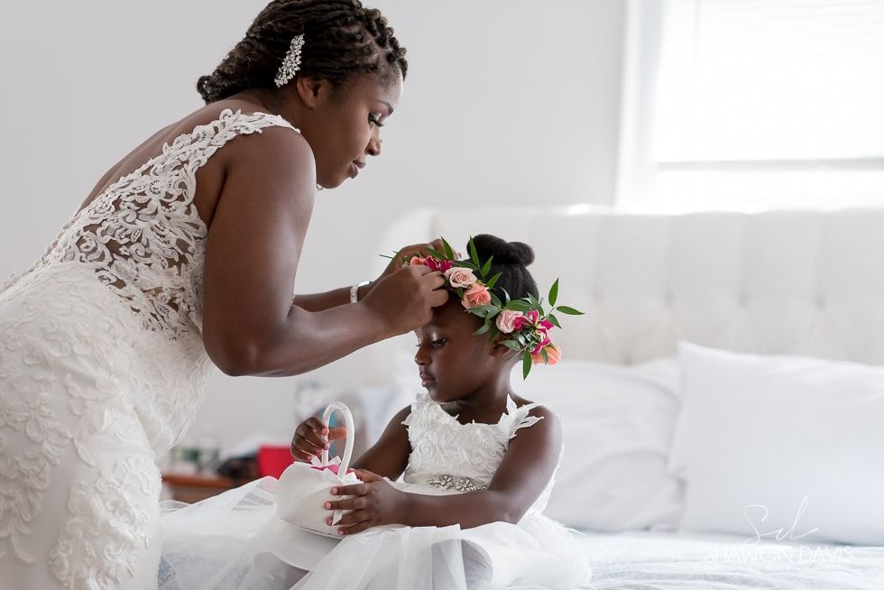 Bride adjusting flower crown on child seated on a white bed