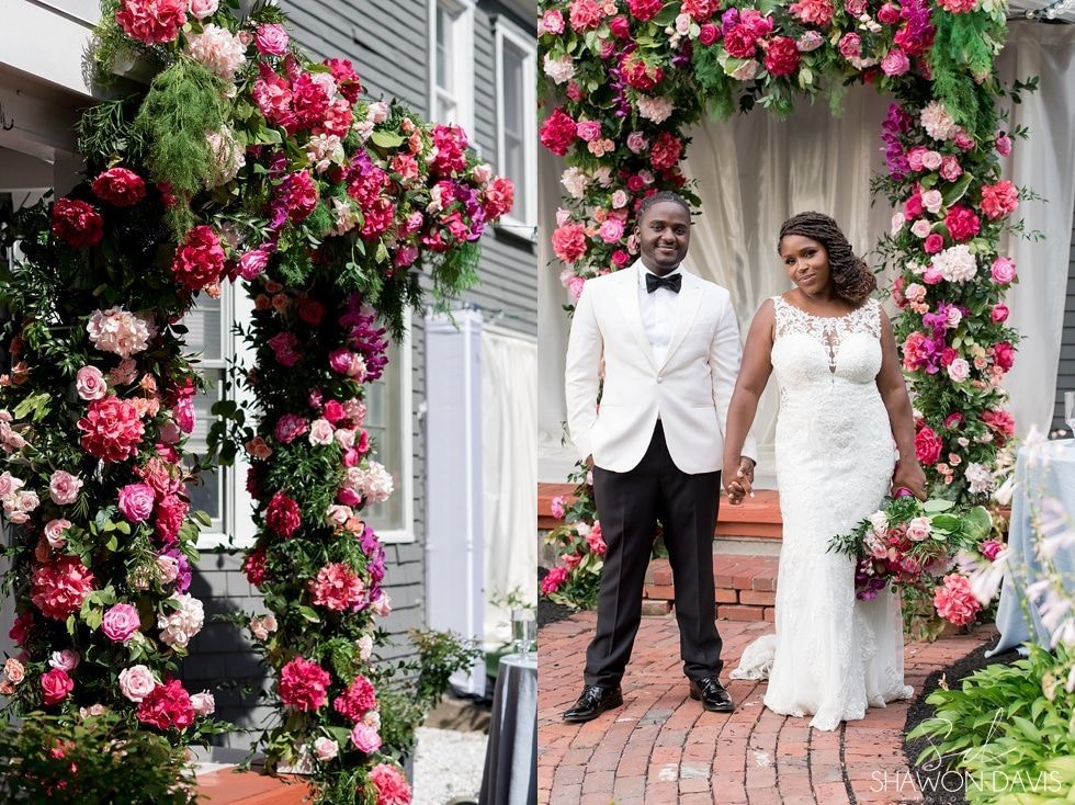 Wedding couple posing beneath pink floral archway on a brick path
