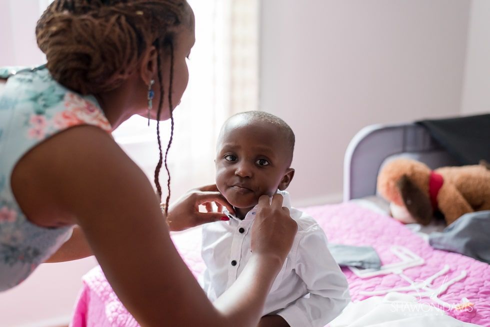 Adult helping a child adjust a bow tie on a bed in a softly lit bedroom