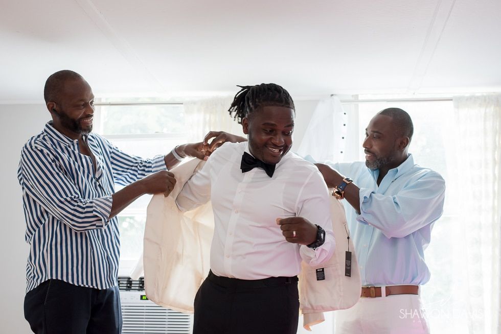 Three men help a man into a light pink suit jacket in a bright room