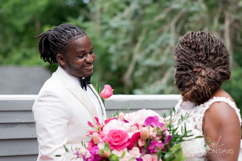 Two people in white outfits smiling during an outdoor wedding, with pink flowers in the foreground