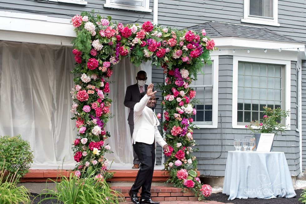 Person stands under a pink floral arch outside a house, holding up a phone during an event.