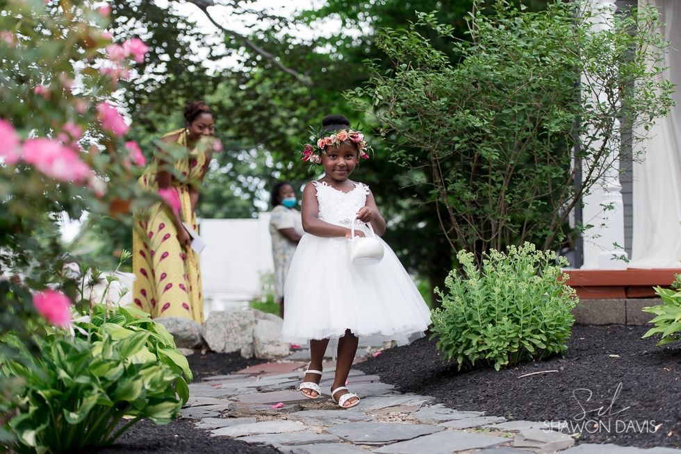 Child in a white dress holding a basket in a garden path, with pink flowers and greenery nearby