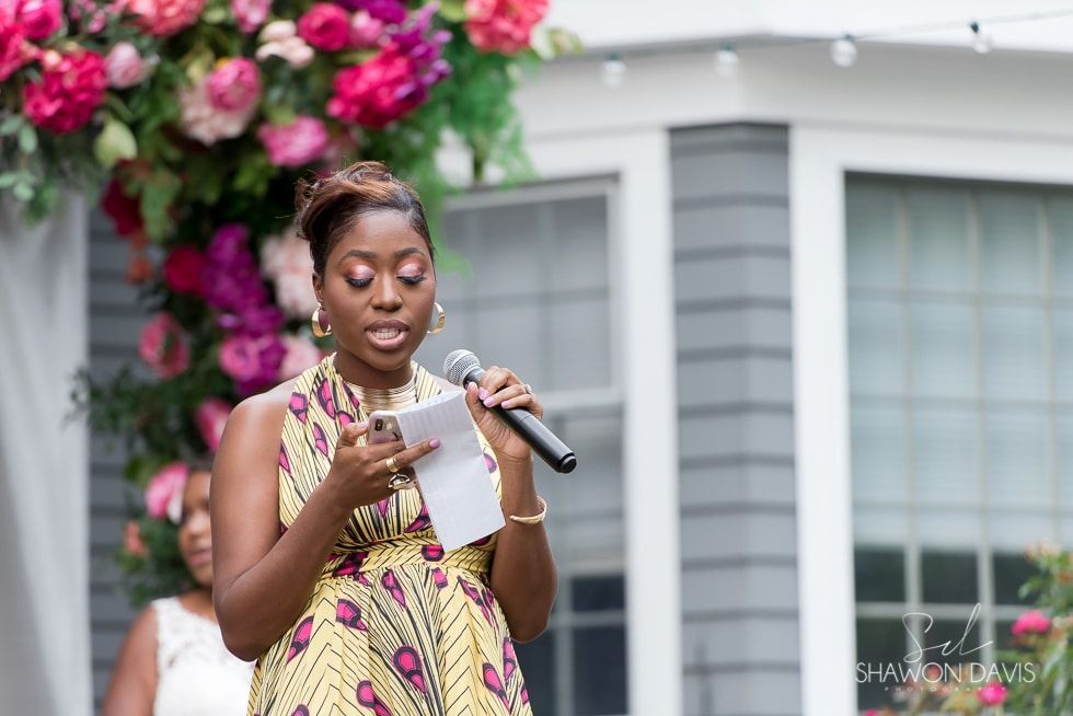 Woman reading from a paper and speaking into a microphone at an outdoor event, with flowers and a house behind her
