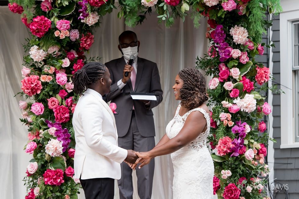 Wedding ceremony outdoors under pink floral arch, couple holding hands as officiant speaks.