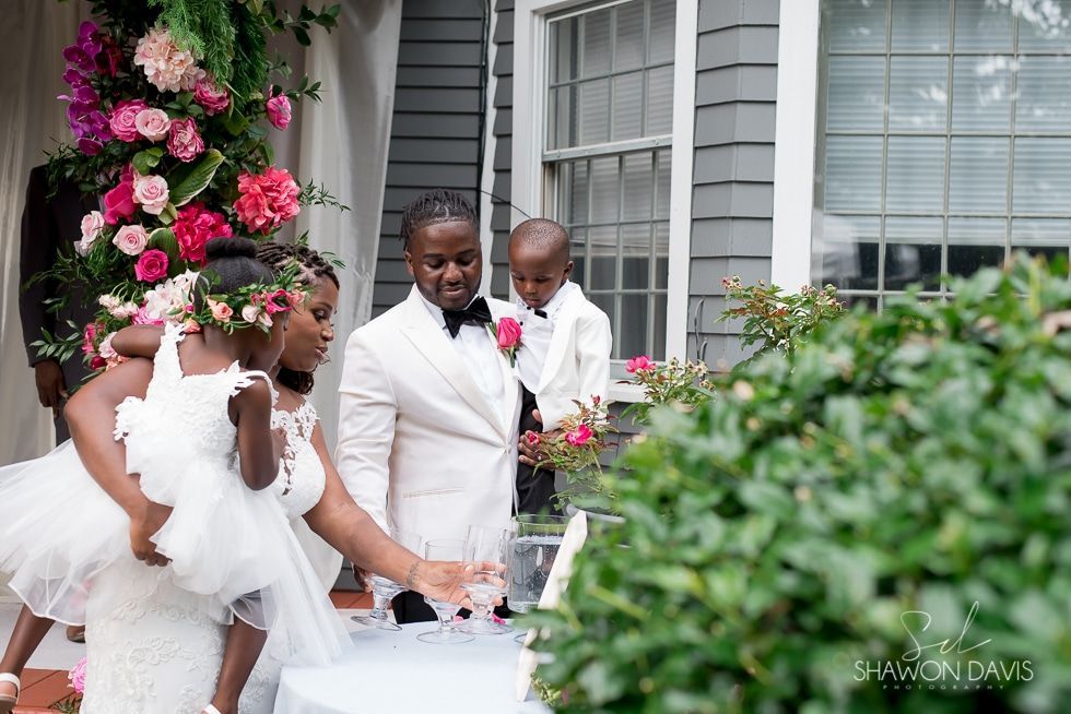 Wedding party in white outfits cutting a cake outdoors beside blooming pink flowers.