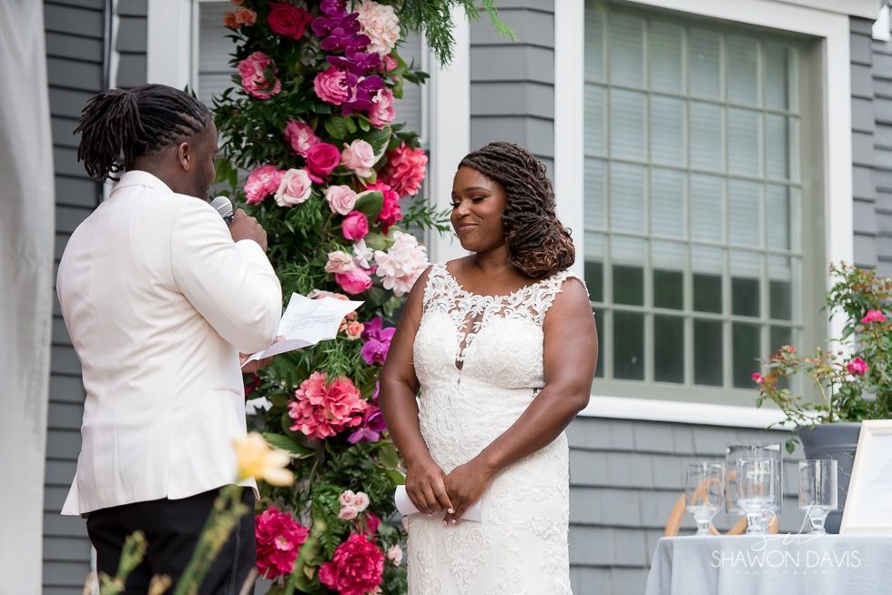 Couple exchanging vows at an outdoor wedding, with pink floral decorations and a decorated table nearby
