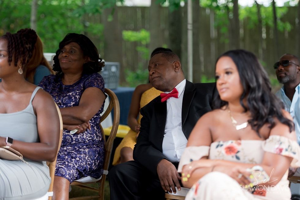 People seated at an outdoor ceremony, including a man in a black suit with a red bow tie.