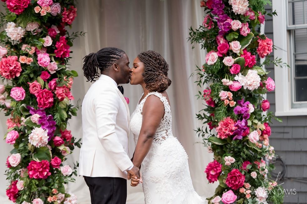 Couple in white wedding attire kissing beneath a pink floral arch outdoors