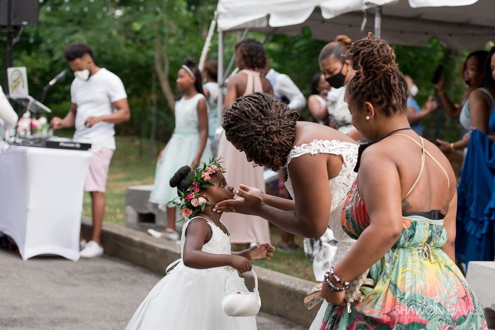 Child in white dress receiving flowers at an outdoor wedding celebration with guests nearby