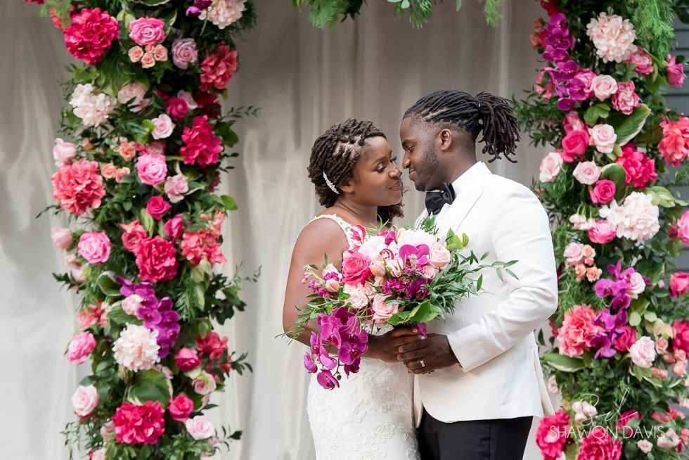 Wedding couple embracing beneath a pink-and-white floral arch, holding a bouquet