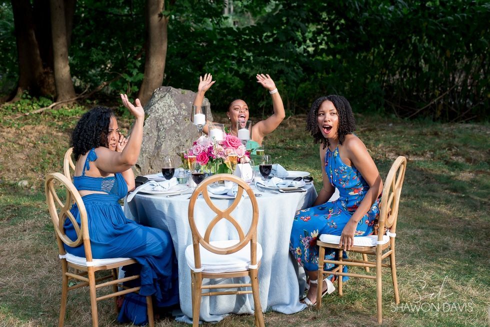 Three women in blue outfits toast at a white-draped outdoor table with flowers and drinks.
