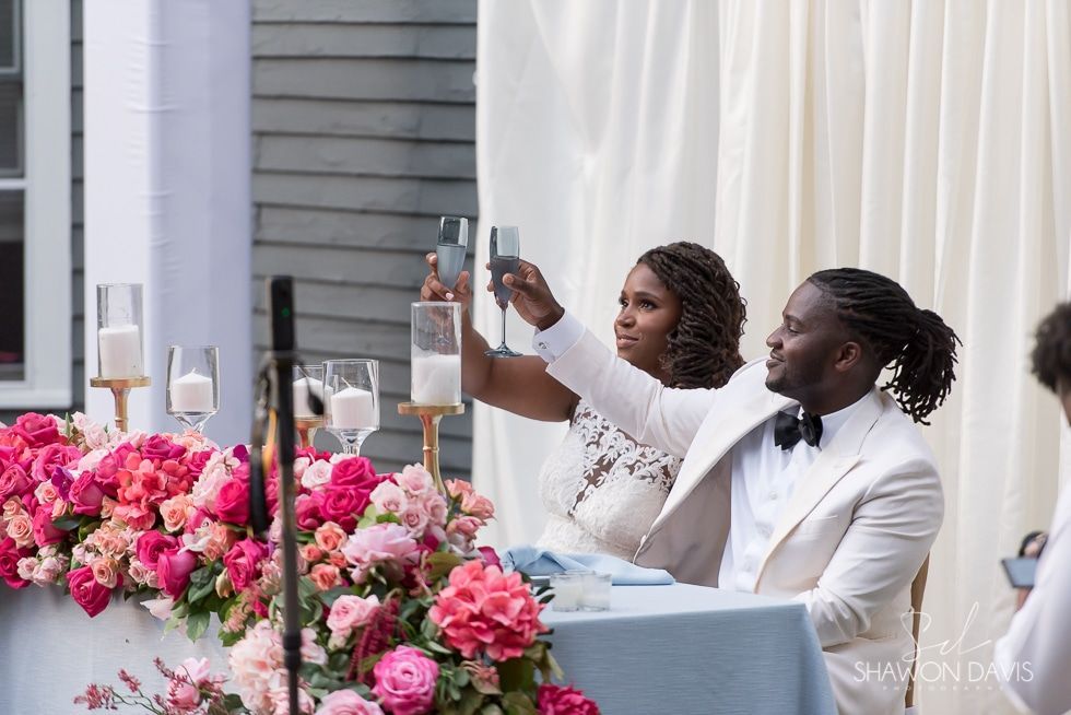 Two people in white suits toast at a decorated table with pink flowers and champagne glasses.