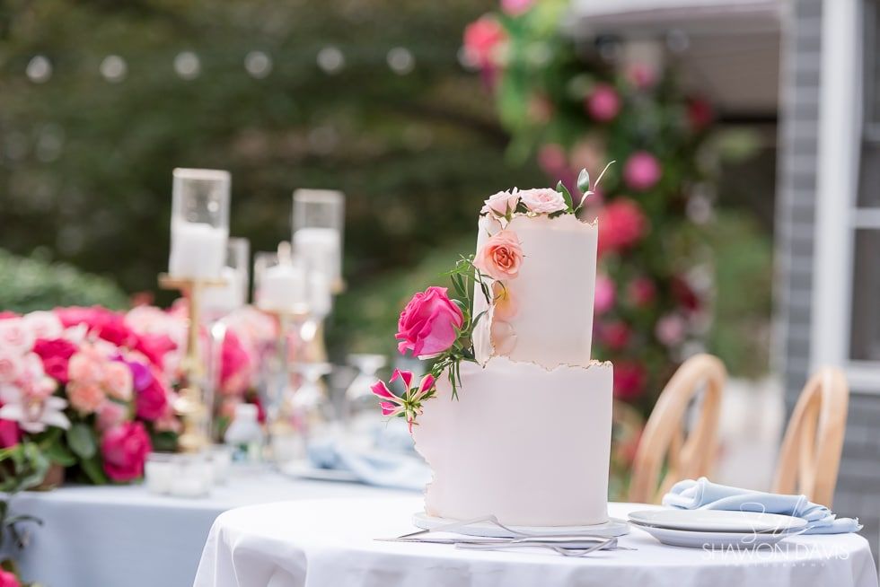 Two-tier white floral cake on a decorated table with pink flowers and candles outdoors