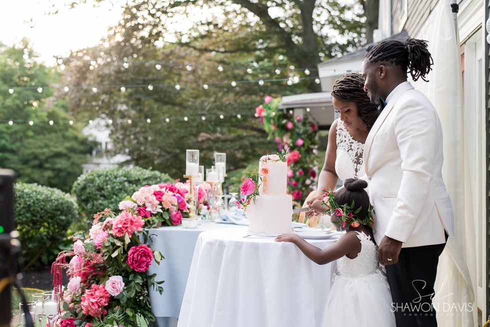 Couple cutting a wedding cake at an outdoor reception with pink flowers and white tablecloths