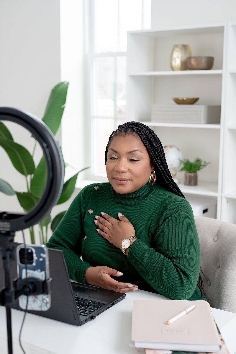 Woman in green sweater speaking on camera at a desk in a bright home office