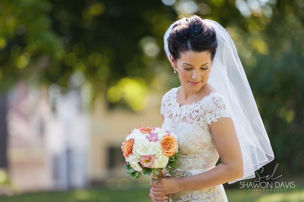 Bride in a white lace dress holding a bouquet, standing outdoors with a soft green background