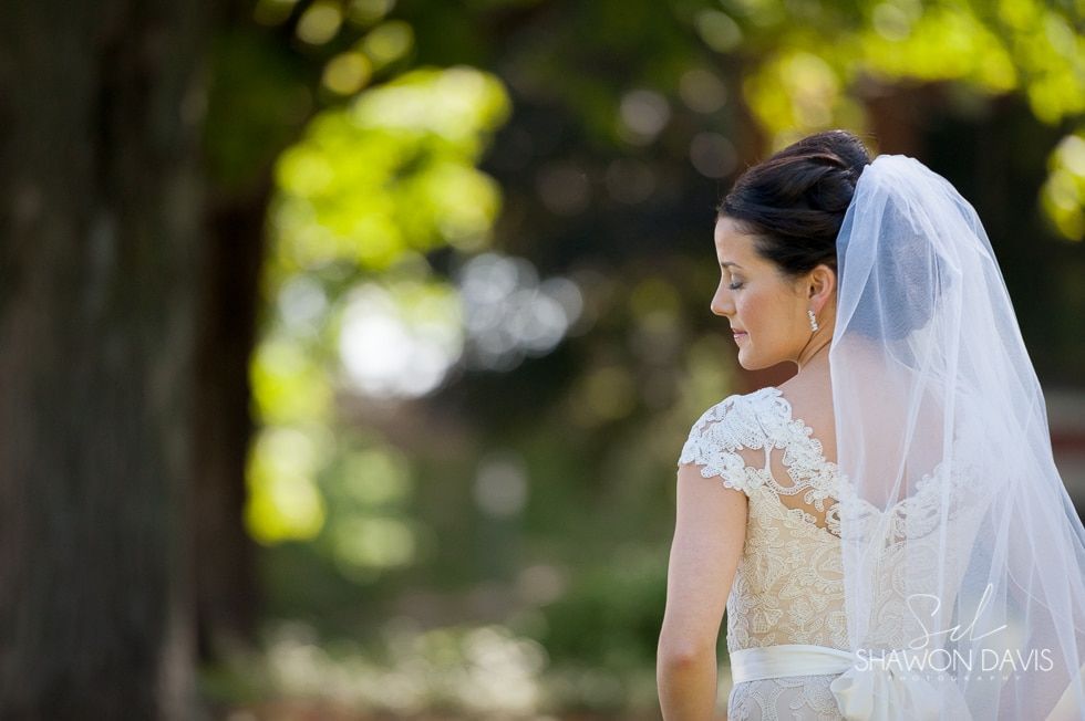 Bride in a white lace dress and veil standing outdoors, turned in profile against a blurred green background