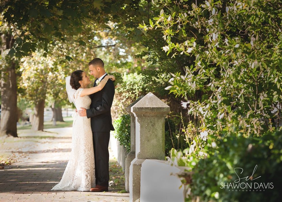 Bride and groom embrace under trees on a sunlit path beside a stone post