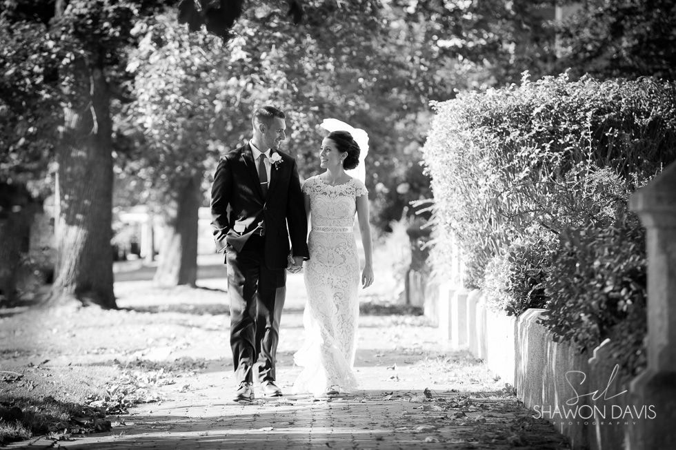 Couple walking arm in arm down a tree-lined path in formal attire, black-and-white photo