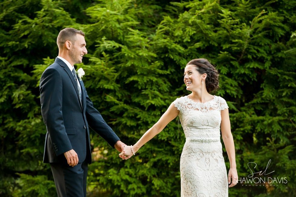 Bride and groom holding hands outdoors in front of green trees, smiling at each other.