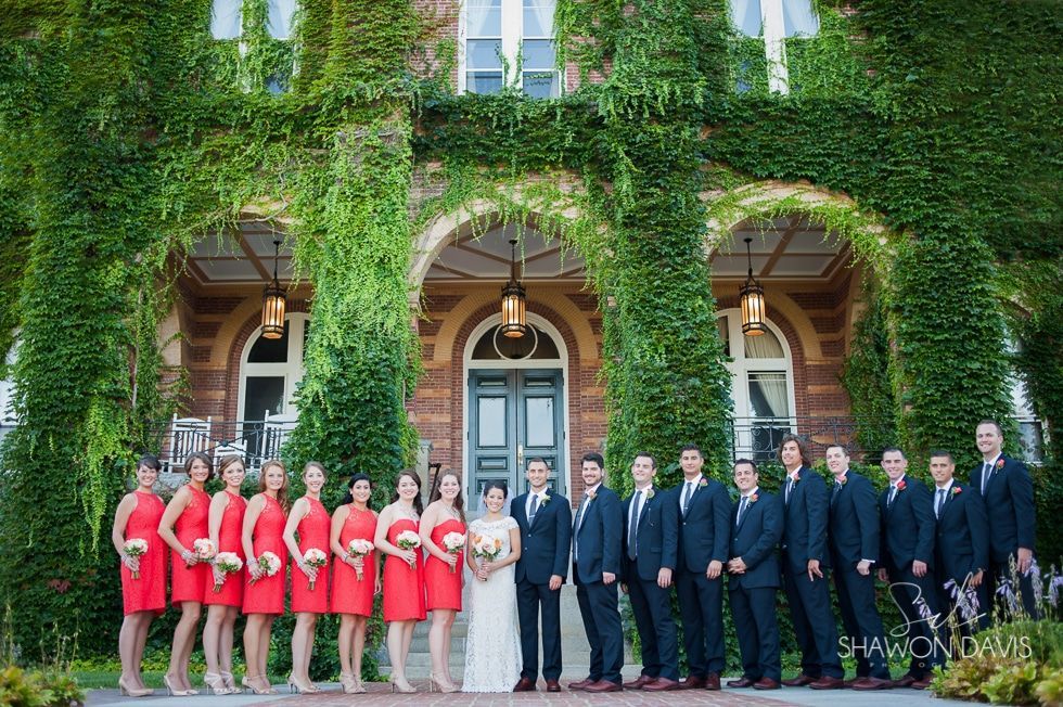 Wedding party posing outside ivy-covered arches of a stone building, bridesmaids in red and groomsmen in navy suits