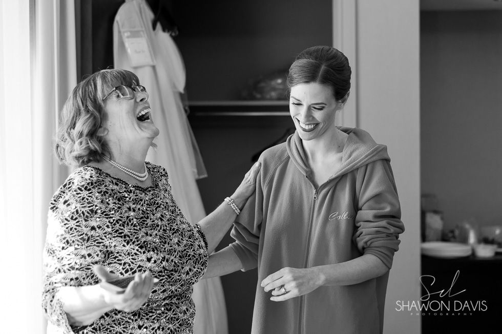 Two women laughing while adjusting sleeves in a dressing room, one holding a cup.